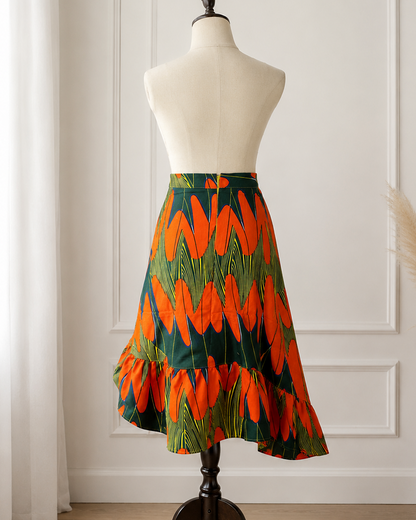 A flared skirt displayed on a cream dress form mannequin in a bright room with white paneled walls, light wood flooring, sheer curtains, and a vase of pampas grass. The skirt features a bold abstract pattern in bright orange and dark green.