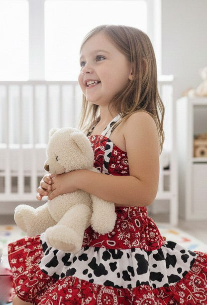 A young girl holding a teddy bear in a room with white walls and furniture, wearing a red, black, and white Western Bandana-patterned and Cow print Crop top.