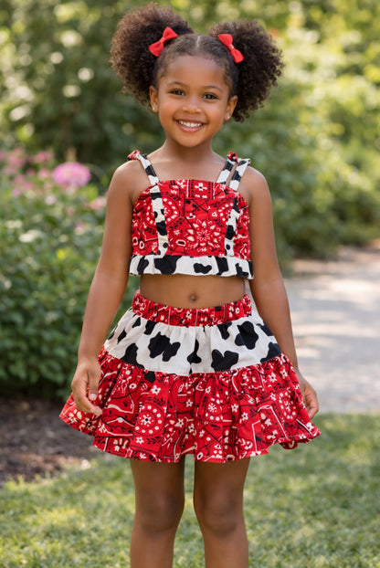 A young girl wearing a red-and-black bandana and a cow-print Skirt and crop top standing outdoors.