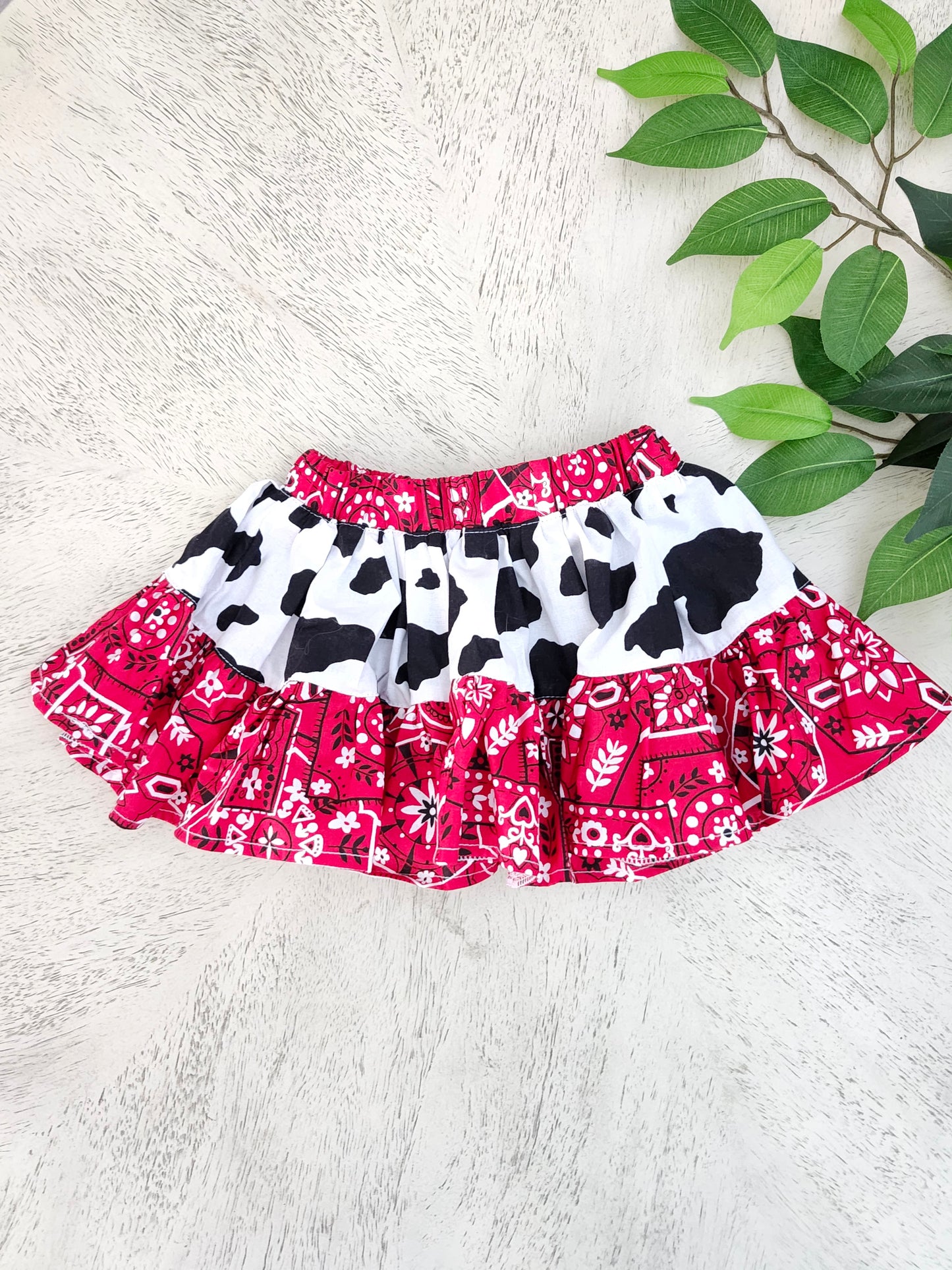 Red, black, and white Western Bandana-patterned and Cow print Tiered Skirt on a wooden surface with green leaves.