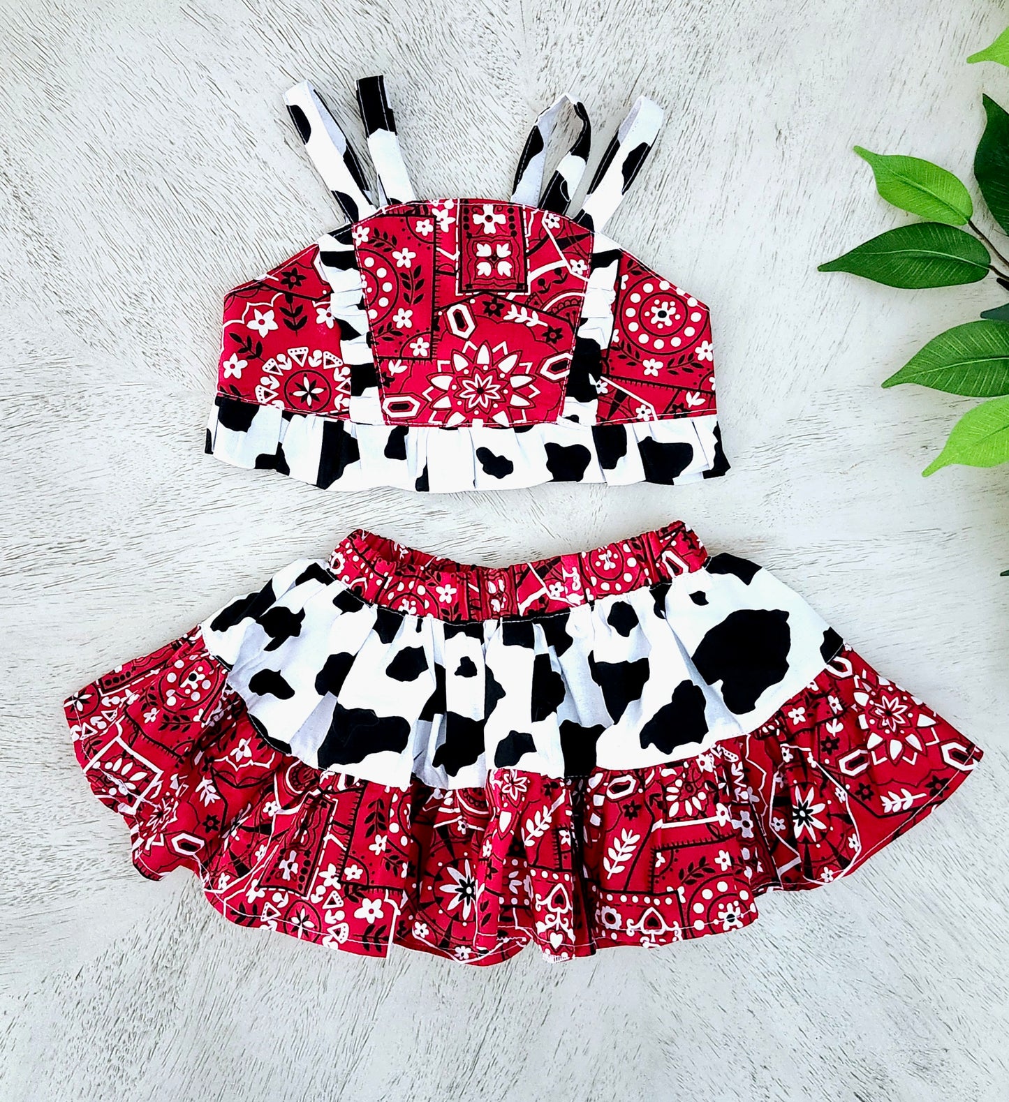Children's outfit with red, black, Bandana Print, and white cow print top and skirt on a light wooden surface.